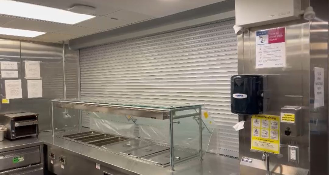 White roll shutter closed across a commercial kitchen servery opening beside stainless steel food service equipment