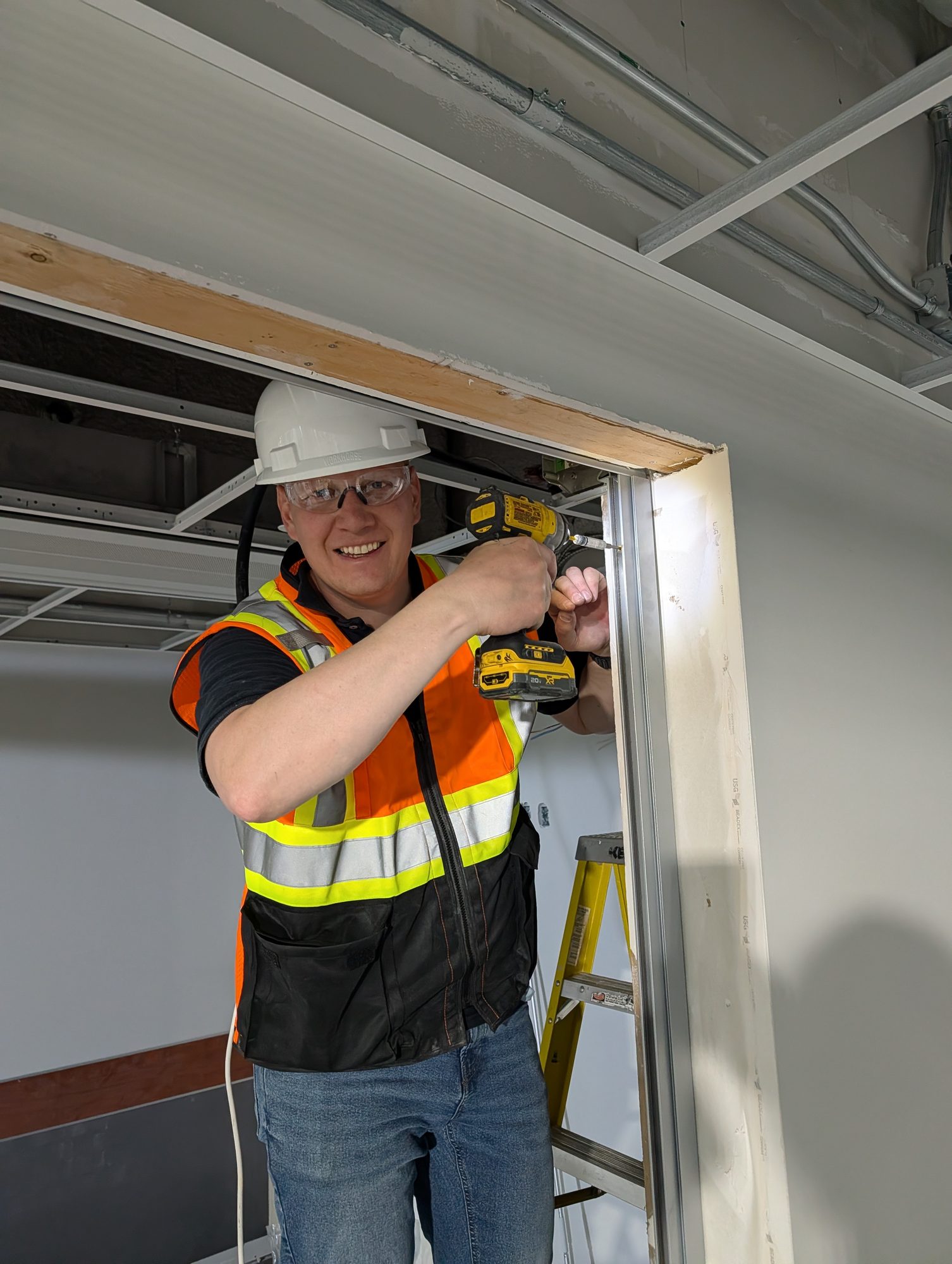 Canada Shutter Pros installer in hard hat and hi-vis drilling a shutter guide rail into a commercial door opening