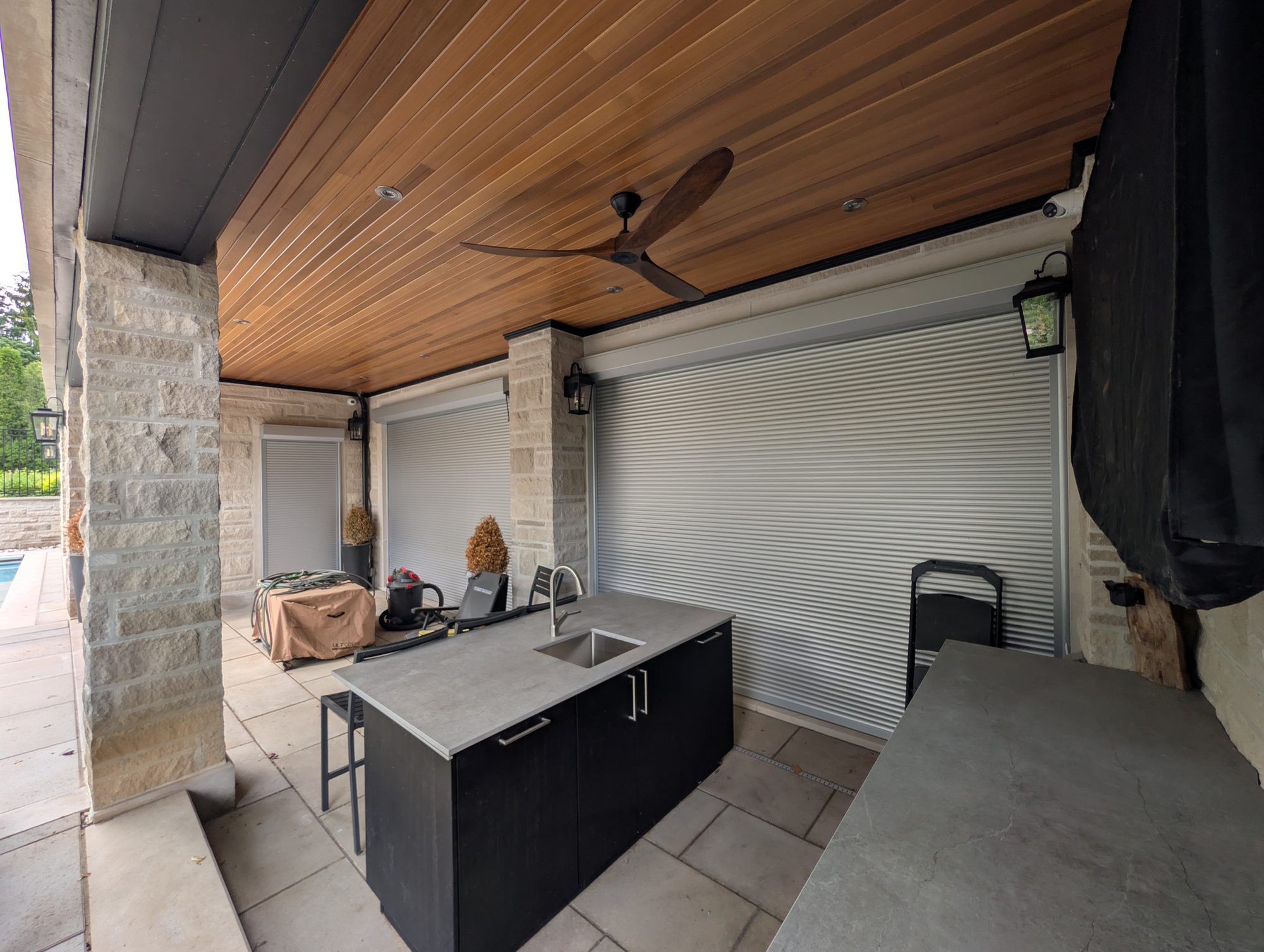 Covered outdoor kitchen with cedar ceiling and roll shutters closed across the rear openings beside a pool