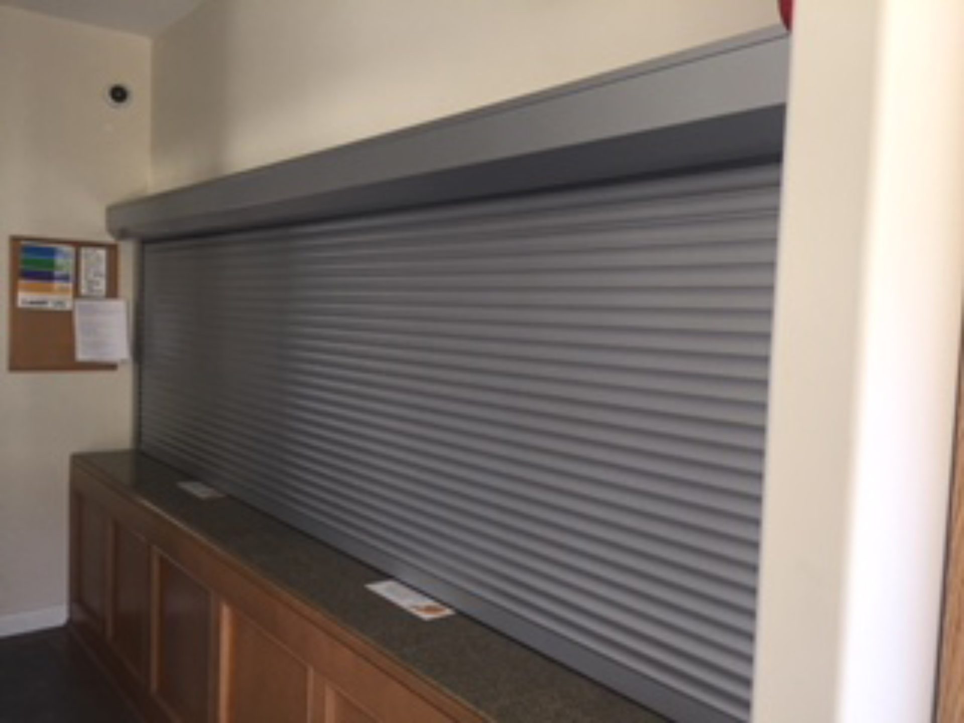 Grey roll shutter closed across a long interior service counter with timber millwork below and a notice board alongside